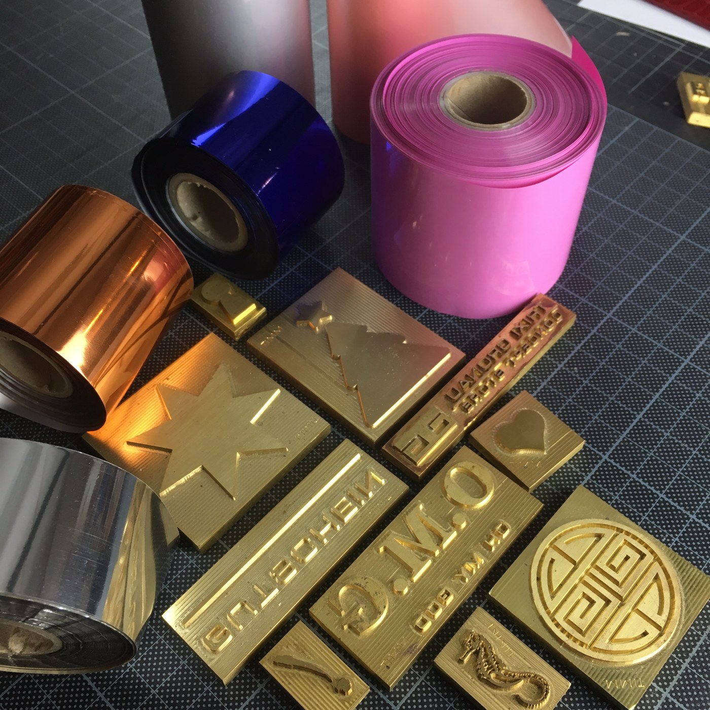 Goldene Messing-Klischees neben Heißfolien-Rollen in Kupfer, Blau, Pink und Silber