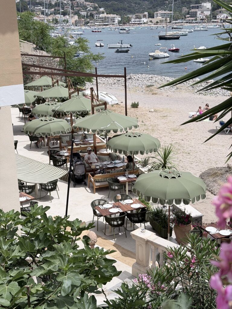 Terrasse des Agapanto in Port de Sóller mit olivgrünen Sonnenschirmen, dahinter die Bucht mit Booten und Strand.