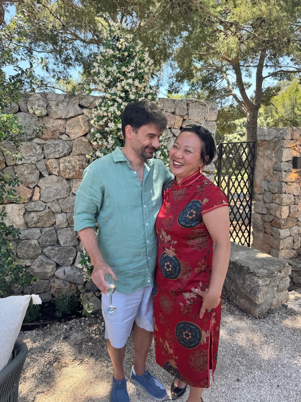 Wing im roten Cheongsam mit Lotusblueten und Juan im salbeigruenen Leinenhemd vor Trockenmauer