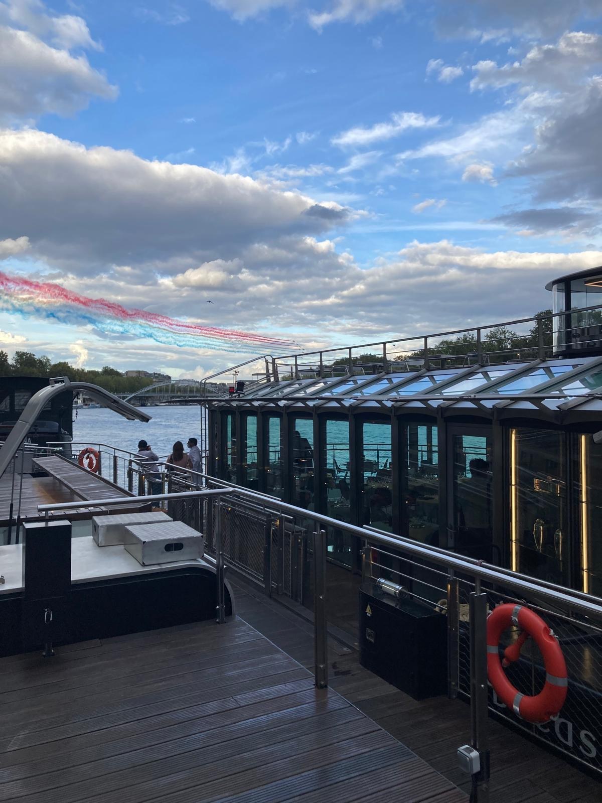 View across the deck of Ducasse sur Seine at the jetty, glass salon and Patrouille de France in the sky