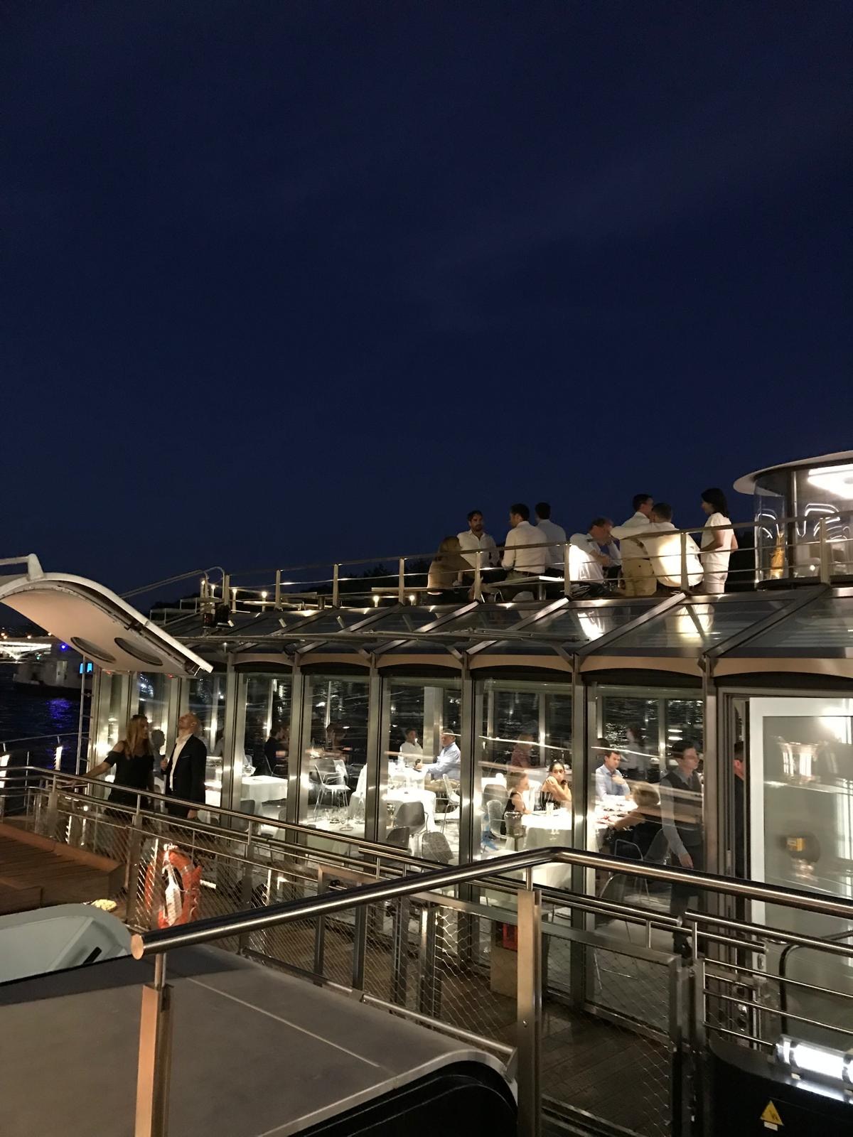 The fully glazed salon of Ducasse sur Seine in the evening, guests on the upper deck