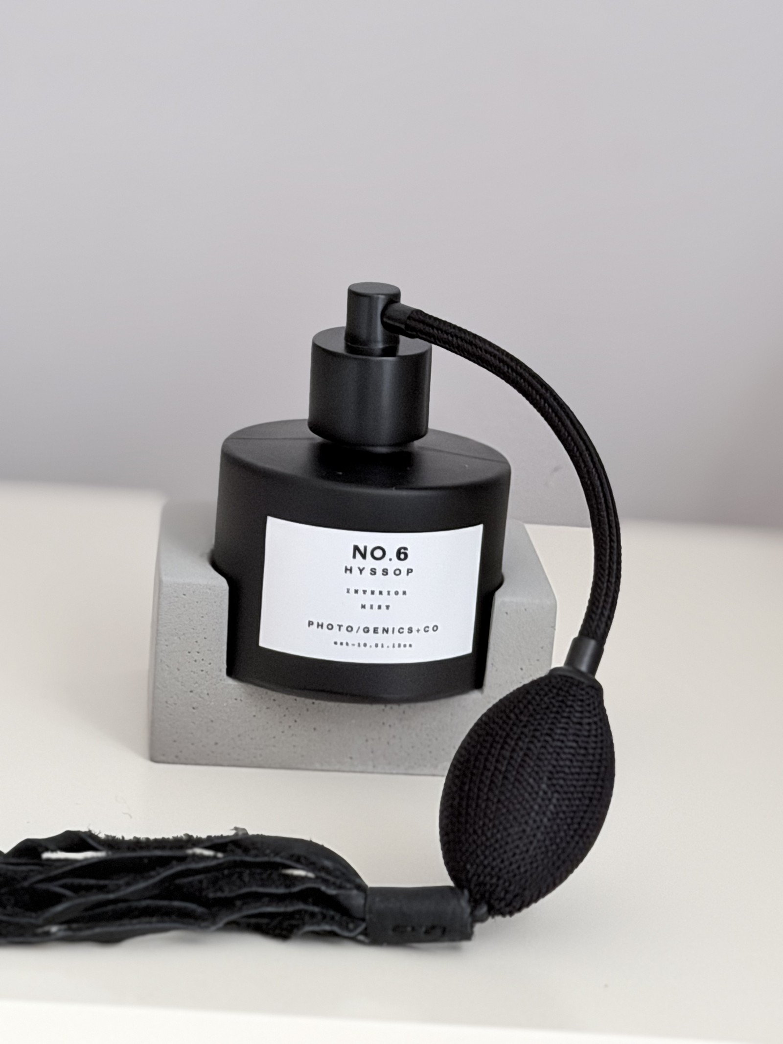 Photo Genics Interior Mist No.6 Hyssop, schwarze Flasche mit Aigrette-Sprayer und Lederquaste auf Betonsockel, Studio-Aufnahme