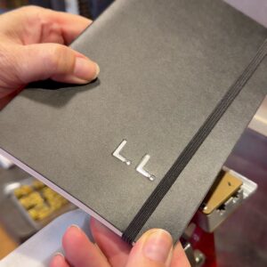 Schwarzes Notizbuch mit silbern geprägten Initialen L.L., in den Händen gehalten im Atelier von Trixi Gronau