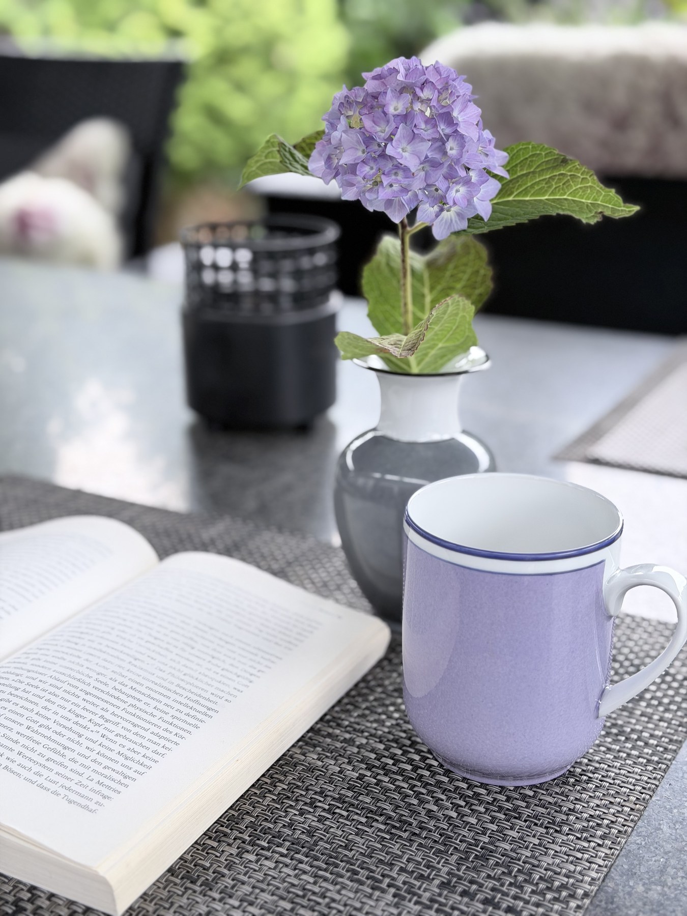 Reichenbach Becher in flieder auf der Terrasse, daneben eine Hortensie in einer grauen Reichenbach-Vase, links ein aufgeschlagenes Buch