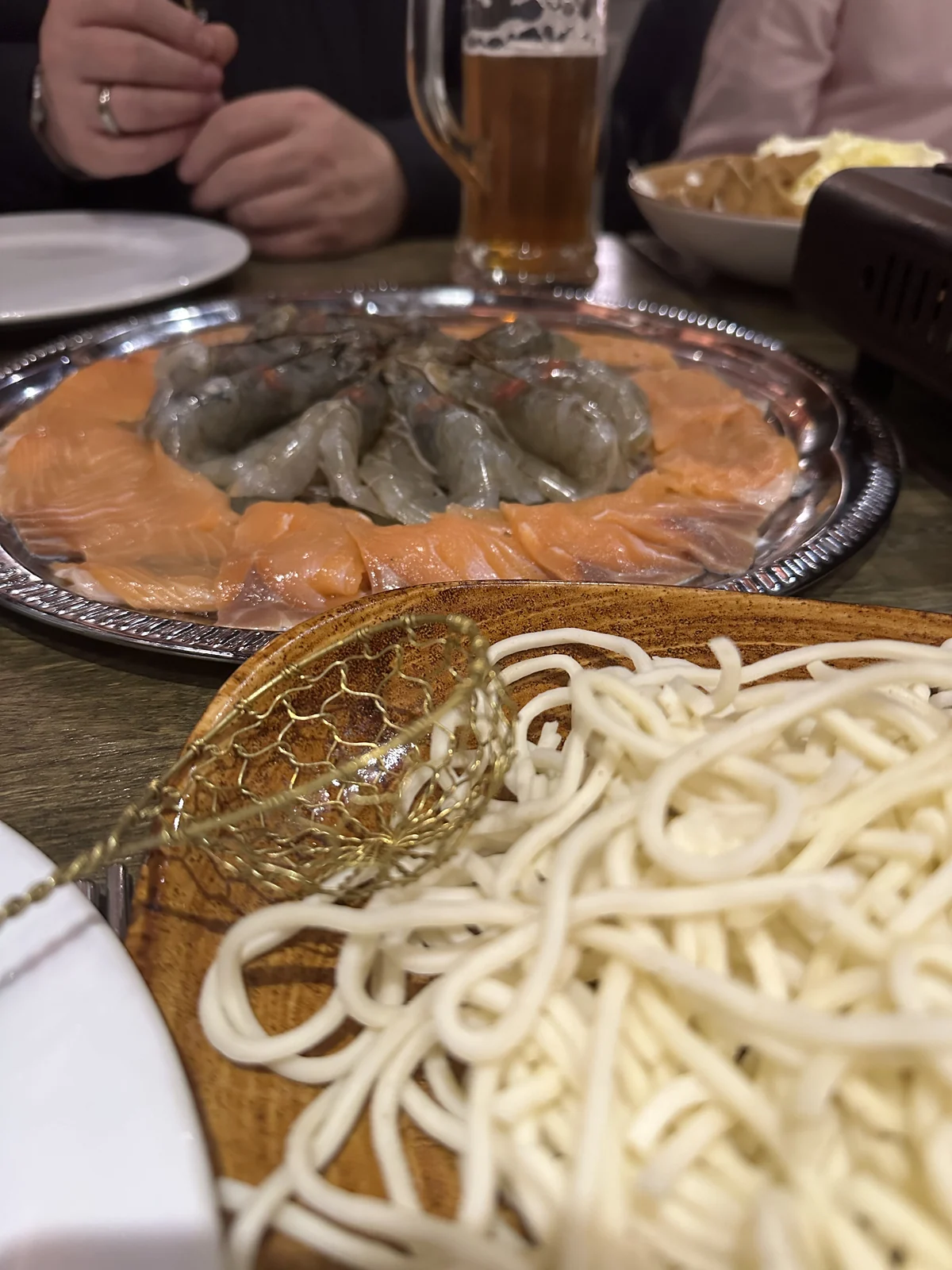 A silver platter of raw prawns and salmon with hand-pulled noodles and a small golden sieve, laid out for Chinese hot pot at Wong restaurant in Hamburg