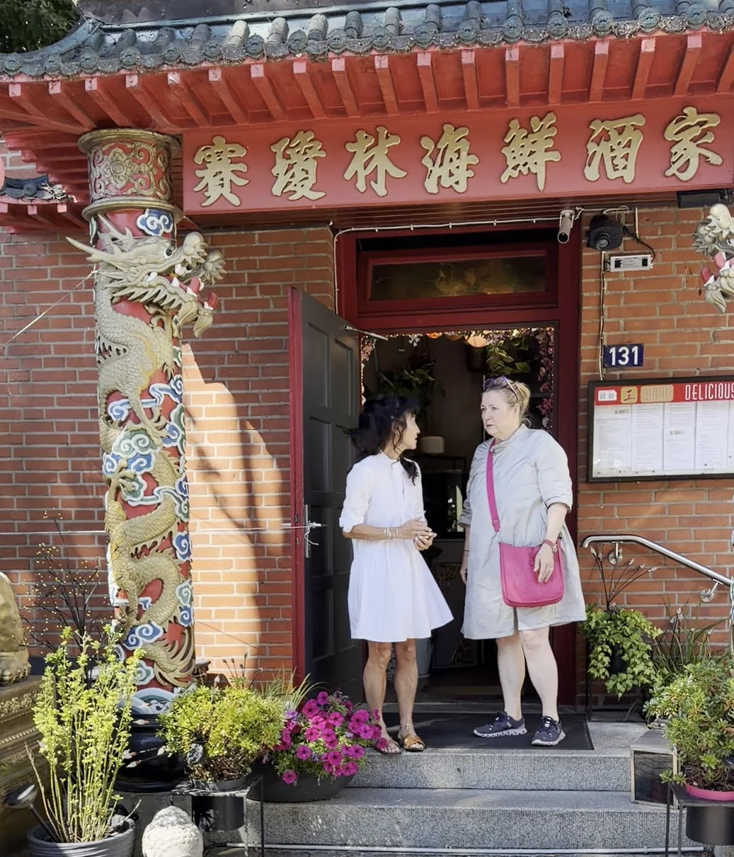 Michelle Wong in a white dress and Trixi Gronau outside the open entrance of Wong restaurant in Hamburg-Langenhorn, beside a gilded dragon column