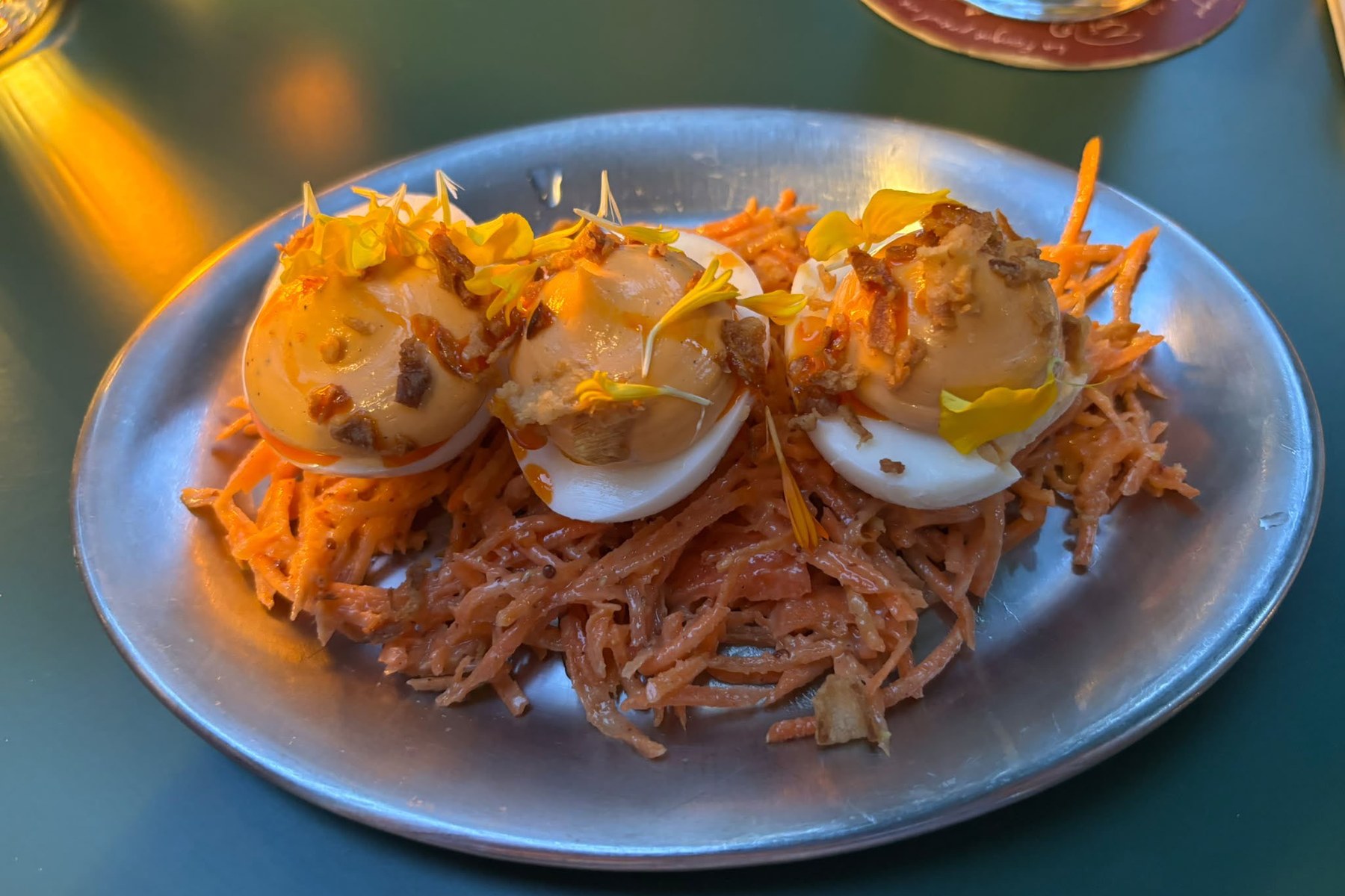 Three halved eggs topped with orange mayonnaise, yellow petals and crisp brown pieces, on a bed of fine orange vegetable strands, on a blue-grey plate