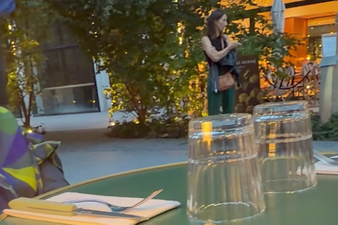 Blick vom Tisch auf der Terrasse: zwei umgedrehte Gläser, eine Serviette und dahinter eine Frau vor der beleuchteten Auslage