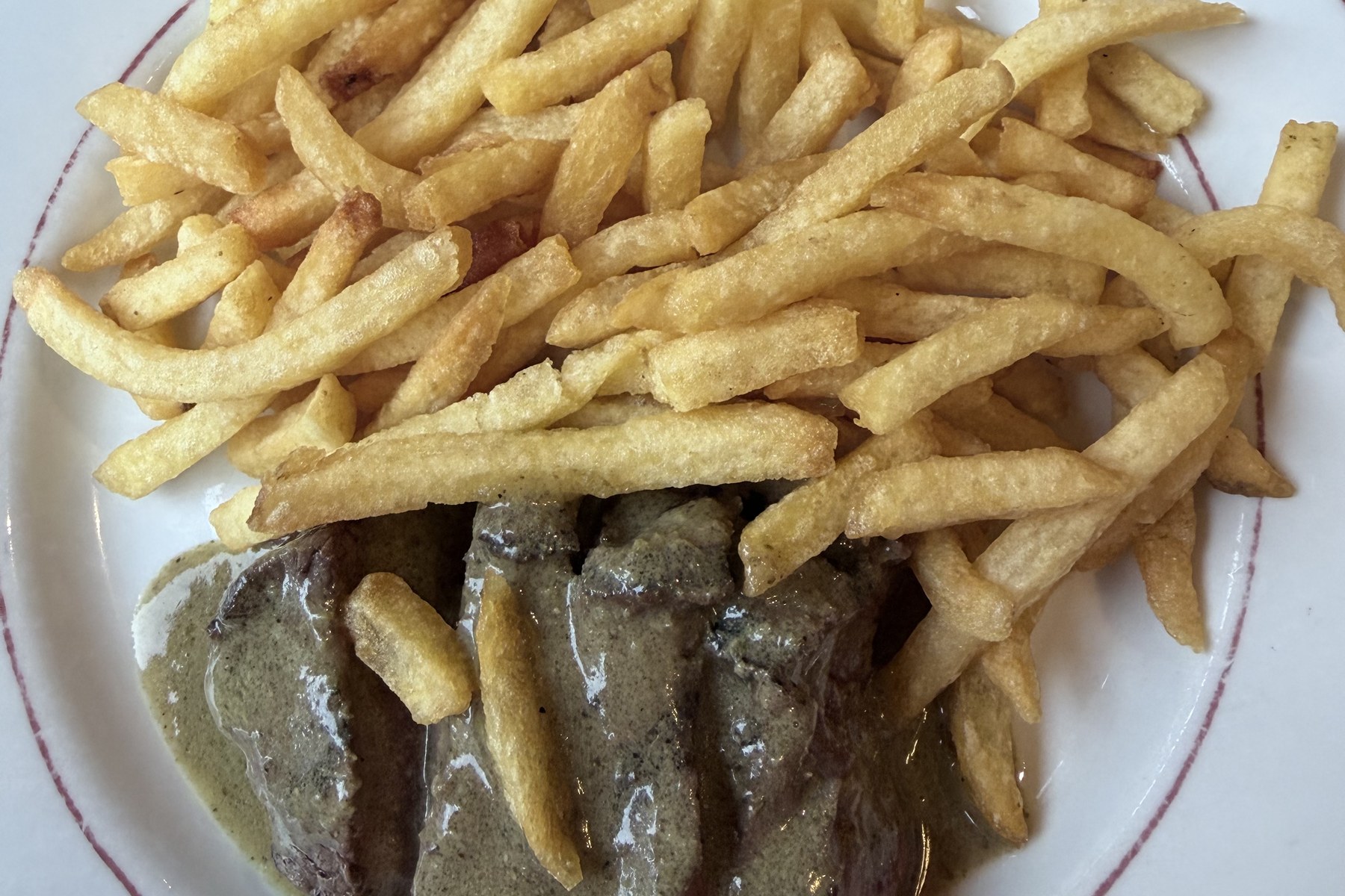 Entrecôte in Scheiben unter einer Portion Pommes allumettes auf einem Teller mit rotem Rand